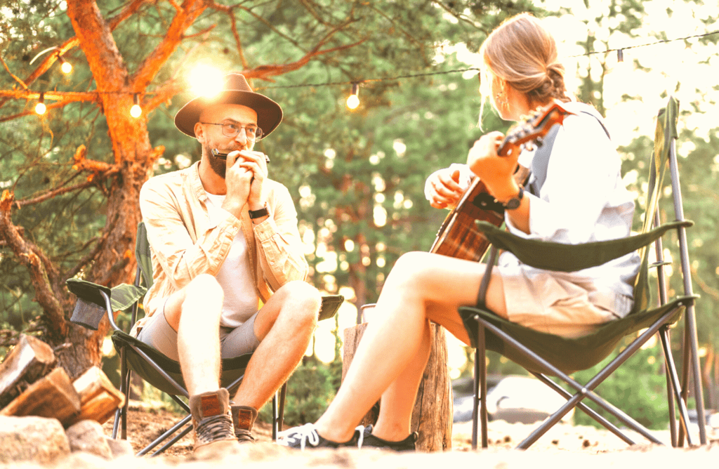 A woman playing the guitar and a man playing the harmonica at the same time on a camping trip